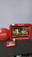 Photo showing the round Coca-Cola frisbee, rectangular TV tray, small rectangular tray, and a white golf ball on top of the TV tray.