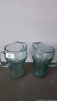 Photo showing two green Coca-Cola glass pitchers side by side with handles and spouts, highlighting embossed Coca-Cola logo and textured glass surface.