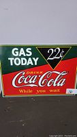 Metal Coca-Cola sign showing gas price 22 cents and slogan "Gas Today, Drink Coca-Cola While you wait" in red, green, yellow and white colors.