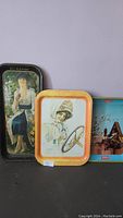 Three Coca-Cola metal serving trays leaning against a wall showing vintage advertising artwork.