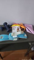 Overview of exercise equipment including blue neoprene hand weights, blue fabric ankle weights, orange resistance band, and packaged Everlast Pilates resistance bands on a table.