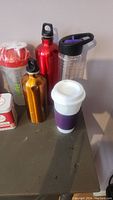 Photo showing five reusable water bottles of different colors and a white reusable coffee cup with purple band