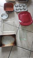 Cookware and bakeware assortment including red microwave-safe dish, metal pans, and glass dishes on tiled floor.