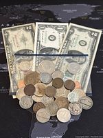 Assortment of coins and three banknotes laid out on a dark map background. Coins include silver and bronze colored items in various conditions. Three banknotes are visible: $2, $1, and $5 bills, showing wear.