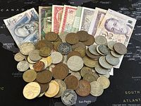 Photo showing numerous assorted world coins scattered with 8 different colored banknotes fanned out behind coins on map background.