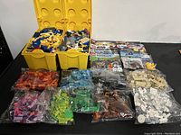Photo showing two yellow storage containers filled with assorted LEGO pieces and multiple bags of sorted LEGO bricks of different colors, with LEGO set boxes visible in the background.