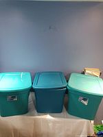 Three teal Sterilite storage bins with lids on a table against a blue wall