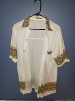 Front view of cream-colored short sleeve Cabana shirt featuring patterned gold and brown accents on collar, cuffs, and hem, hanging on black hanger against blue wall.