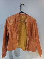 Front view of brown leather look jacket hanging on black hanger against a light gray wall.