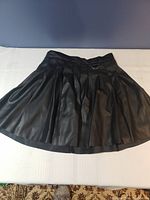 Front view of a black pleated faux leather skirt with a metal D-ring detail on the waistband.