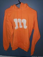 Front view of bright orange hoodie with large white M on chest, hanging on black hanger against blue background.
