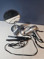 Photo of vanity mirror, Revlon hair dryer, black hair straightener, and another hair styling tool all placed on a white surface against a blue wall.