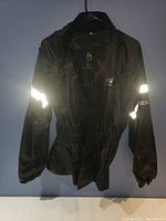 Front view of black rain jacket with reflective stripes on sleeves and elastic waistband, hanging on a hanger.