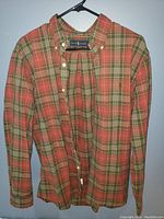 Front view of the plaid button-up shirt hanging on a black hanger on a white wall, showing red, green, and beige plaid design.