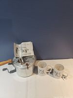 Aluminum stovetop popcorn popper with wooden handle, two white ceramic mugs labeled 'MR' and 'MRS', and instruction manual placed inside the popcorn popper lid.
