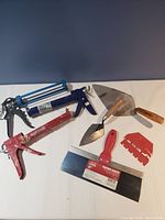 Overview of 5 construction tools including caulking guns, masonry trowels, taping knife and multi-notch drywall tool on white surface.