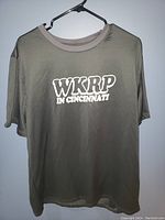 Front view of gray WKRP in Cincinnati T-shirt hanging on hanger against wall.