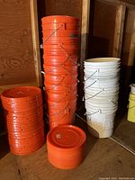Stack of orange and white 5-gallon buckets with metal handles, stacks of orange lids next to buckets.