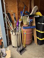 View of two large barrels filled with long-handled cleaning tools including brooms, swiffer, and a dustpan, plus two sledgehammers leaning against a barrel.