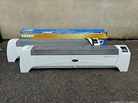 Two Lasko baseboard heaters with white plastic ends and metal grates, one in original box, placed on the ground against a wall.