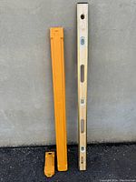 Full view of the 48 inch wooden Johnson spirit level alongside its yellow protective hard case placed against a concrete wall.