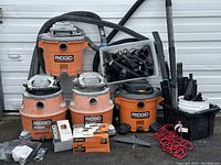 Four Ridgid Blower Vacuums and multiple accessories arranged outside a garage door including hoses, fittings, a red extension cord, and two black plastic containers holding additional attachments and parts.