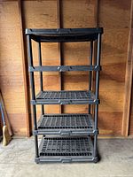 Front view of the assembled five-tier black plastic shelving unit against a wood wall, showing all shelves and structure.