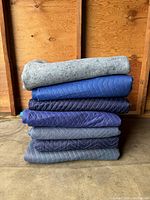 Stack of seven moving blankets, six quilted in shades of blue and gray, one gray felt blanket on top, folded and stacked against a wood wall.