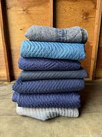 Stack of seven moving blankets showing different textures, colors, and material types.