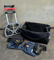 Magna Cart collapsible two wheel cart with rubber tires, two fabric tool belts, and black duffel bag on ground.
