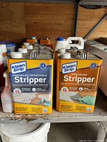 Two Klean Strip premium stripper metal jugs, one sprayable liquid formula (blue label) and one paste formula (orange label), side by side on shelf.