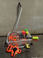 All items arranged in plastic bin on ground against wall showing measuring wheel, flags, and spray cans