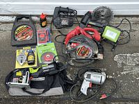 Full lot of power tools and accessories laid out on pavement, showing Rockwell Versacut saw, SKIL circular saw, Black & Decker jig saw, Stanley electric stapler, LED light, DeWalt chalkline kit, wire brush, extra saw blades, multimeter, and Craftsman drill.