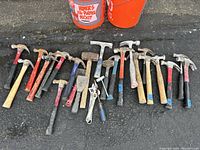 Photo showing 21 assorted hammers of varying sizes and head types arranged on ground with two orange plastic 5-gallon buckets in the background.
