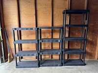 Three black plastic shelving units lined up side by side against a wooden wall in a garage or storage space. Two units have four shelves each and one unit has five shelves.