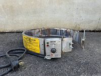 Wide shot of the WRAP-IT-HEAT metal band heater with cord and control panel, showing rust and wear, on asphalt ground.