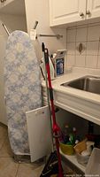 Photo of ironing board, broom, and mop near countertop and sink in kitchen or utility space.