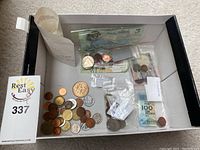 Wide view of box containing various coins and paper currency, some in ziploc bags. Contains loose coins spread out, a Portuguese banknote, and a labeled bag.