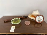 Photo showing carved wooden bat, yellow-green coloured glass bowl with a lattice rim, small pink glass shell dish, large sea shell, and framed round oriental art piece on a wooden surface.