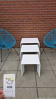Front view of three white nesting tables arranged stacked inside each other outdoors on patio with two blue chairs and brick wall background.
