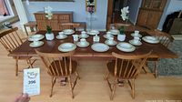 Wide photo of the wooden dining table with six chairs arranged around it, showing the table set with white china dishes and tabletop decor, leaves inserted to extend length.