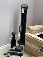 Black tower fan positioned beside sofa, with handheld steamer, LED light, and two power bars on the floor in corner against wall.