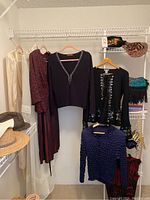 Women's clothing including white dress, maroon dress, black cardigan, black top with sequins, and blue sweater on hangers in closet with hangers and shelving.