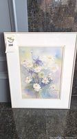 Full view of framed watercolor painting portraying a flower bouquet including daisies and purple flowers, placed against a reflective wall.