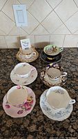 Six Royal Albert china teacups and saucers displayed on granite countertop, showing each cup on its matching saucer in varied floral and decorative designs.