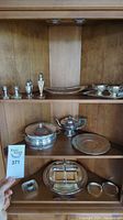 Full view of all silver plated serving dishes spread on wooden shelves including lidded bowls, teapot, trays, condiment holders, and shakers.