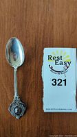 Full view of the sterling silver spoon with detailed crest and emblem near handle end, placed next to a lot number card for scale.