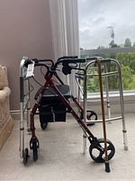 Red Elite rollator walker, silver folding walker, and wooden cane arranged near a window.