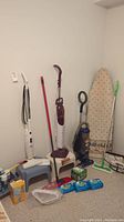 Various cleaning supplies including vacuum cleaners, steam mop, ironing board, step stools, and mop in a room corner.