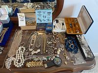 Full view of assortment of costume jewelry spread on wooden surface with a jewelry box and additional items in background
